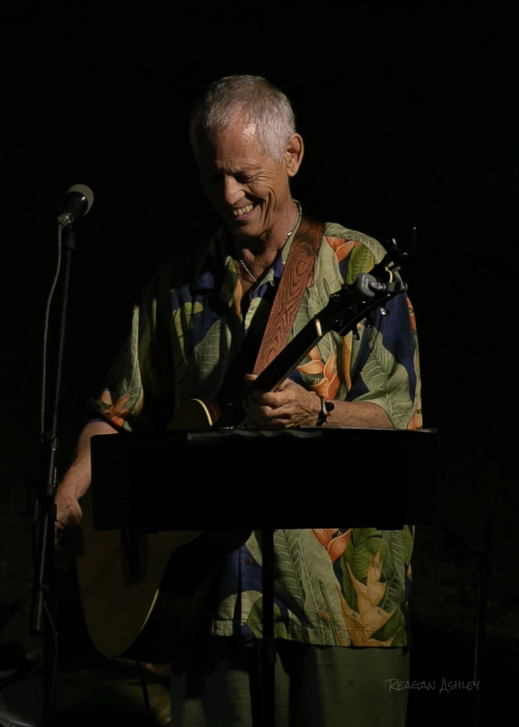 Rudi Harst smiles while holding a guitar on a dark stage with a microphone and music stand.